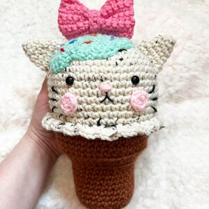Puede incluir: Un cono de helado con forma de gato hecho a mano. El gato tiene un lazo rosa, helado de menta con chispas y mejillas rosas. El cono es marrón. El texto "pattern" está en la parte inferior.