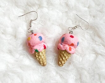 May include: Novelty earrings featuring pink ice cream cones with colourful sprinkles. The ice cream sits on a textured, golden-brown cone. The earrings have silver-coloured fish hook closures, ideal for adding a touch of fun to any outfit.