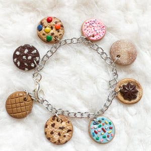 Peut inclure: Un bracelet à chaîne argentée orné de huit breloques en forme de biscuits. Les breloques sont décorées pour ressembler à différents types de biscuits, avec des pépites de chocolat, des vermicelles et des bonbons. Le bracelet est présenté sur un fond blanc et moelleux.