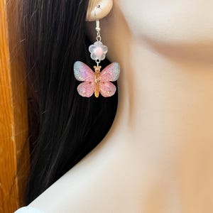 May include: A pair of dangle earrings featuring a butterfly and flower design. The butterfly has pink and blue iridescent wings with a gold-colored body. A small white flower with a pink center is above the butterfly.