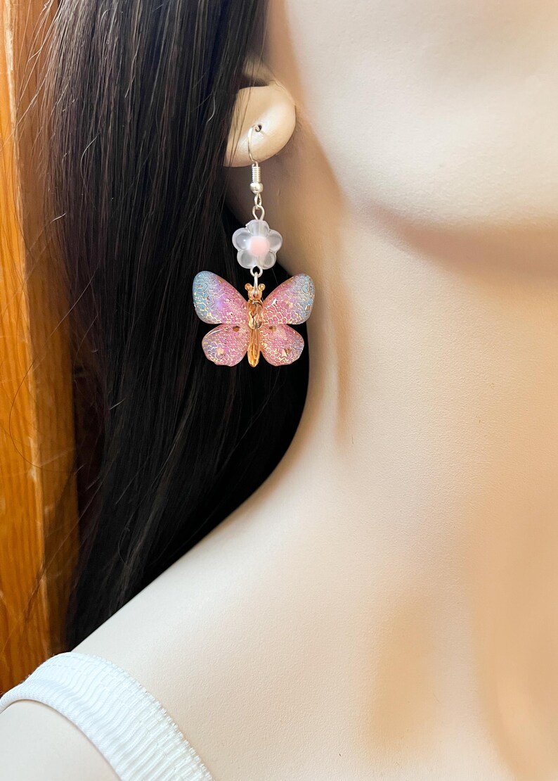 May include: A pair of dangle earrings featuring a butterfly and flower design. The butterfly has pink and blue iridescent wings with a gold-colored body. A small white flower with a pink center is above the butterfly.