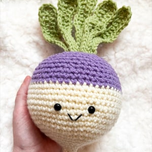 May include: A crocheted turnip toy with a smiling face. The top of the turnip is purple, the body is off-white, and the leaves are green. The word "pattern" is written in the bottom right corner.