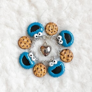 May include: A silver bracelet featuring blue Cookie Monster charms with white eyes and black mouths, alternating with chocolate chip cookie charms. A silver heart charm hangs in the center. The bracelet is set against a white, fluffy background.