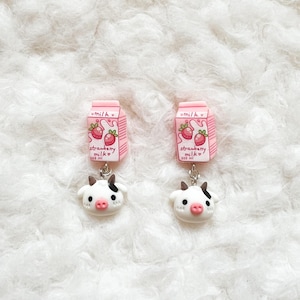 May include: Dangle earrings with pink strawberry milk carton charms and cow head charms. The milk cartons have strawberry illustrations and the text "strawberry milk". The cow heads are white with black spots and pink noses.