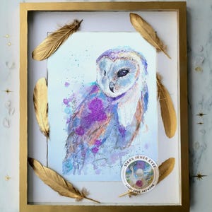 May include: A framed watercolour painting of a barn owl featuring purple, blue, and orange splatters. The artwork is enclosed in a gold frame decorated with gold feathers. A circular sticker in the bottom right corner reads STARS IN HER EYES LIGHT CODE ARTWORK.