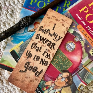 Puede incluir: Una varita de madera oscura descansa sobre dos libros de Harry Potter y un marcapáginas. El marcapáginas tiene el texto "I solemnly swear that I'm up to no good" y huellas. Los libros son rojos y azules.