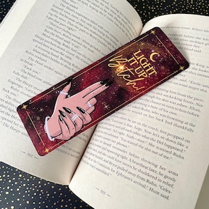 Può includere: Un segnalibro bordeaux con la scritta "LIGHT IT UP Bitch!" presenta un'illustrazione di una mano con unghie lunghe nere e anelli. Il segnalibro è appoggiato su un libro aperto.