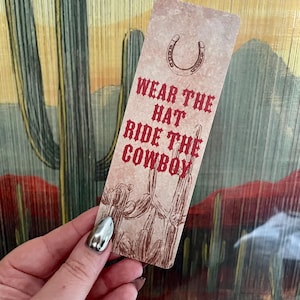 May include: A rectangular bookmark with a beige, textured background featuring a brown horseshoe icon at the top and a brown line-art illustration of a cactus at the bottom. The text WEAR THE HAT RIDE THE COWBOY is printed in red, bold, western-style font.