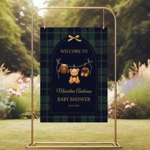 May include: A gold metal stand displaying a navy blue and green plaid baby shower sign. The sign reads "WELCOME TO" above a clothesline with a sweater, teddy bear, and shoes. The sign also includes the name "Marceline Anderson" and "BABY SHOWER".
