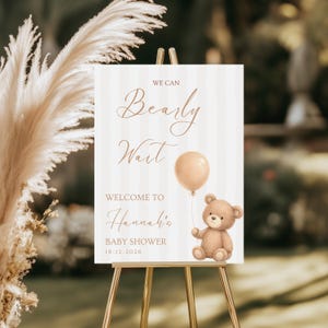 Puede incluir: Un letrero de bienvenida para baby shower sobre un caballete dorado. El letrero tiene un fondo a rayas con las palabras "We Can Bearly Wait" en una elegante escritura. Se incluye una ilustración de un oso de peluche y un globo. El letrero dice "Welcome to Hannah's Baby Shower 16-12-2026."