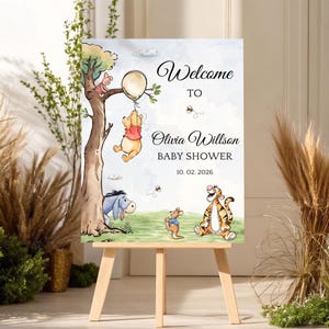 Op de afbeelding: Een welkomstbord voor een babyshower met Winnie de Poeh-personages. Het bord bevat de tekst "Welcome to Olivia Willson BABY SHOWER 10.02.2026" met illustraties van Poeh, Knorretje, Iejoor, Tijger en Kanga. Het bord wordt gepresenteerd op een houten ezel.