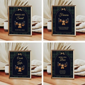 May include: Four framed signs with gold trim, featuring a dark green and navy plaid background. Each sign has a different message in gold script, with a teddy bear and baby clothes graphic. The signs are for a baby shower.
