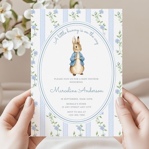 Op de afbeelding: Een uitnodiging voor een babyshower met een aquarel illustratie van een konijn met een blauw jasje. De uitnodiging heeft een blauw-wit gestreepte achtergrond met bloemaccenten en de tekst "A little bunny is on the way."