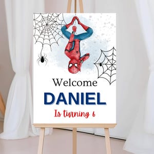 May include: A white sign on an easel featuring a watercolor illustration of Spider-Man hanging upside down. The sign reads "Welcome DANIEL Is turning 6" in blue and red text, with spiderwebs and spiders.