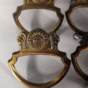 以下が含まれることがあります： アンティーク真鍮製の引き出し取っ手のクローズアップ。各取っ手は上部に装飾的なサンバーストのデザインと湾曲したハンドルを備えています。取っ手はヴィンテージで、古くなった外観をしています。