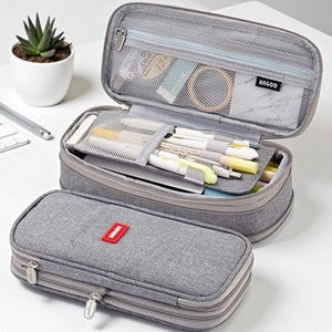 Puede incluir: Estuche gris con cierre de cremallera, abierto para revelar bolígrafos, lápices y artículos de papelería. El compartimento superior tiene un bolsillo de malla. El estuche cerrado tiene una etiqueta rectangular roja. La marca "ANGOO" es visible.