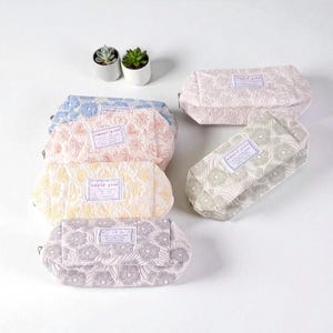Op de afbeelding: Een verzameling cosmetische tassen met bloemmotieven in verschillende kleuren, waaronder roze, blauw, geel en grijs. Elke tas heeft een rechthoekig label met de tekst "sweet girl". Twee kleine vetplanten in potten zijn zichtbaar.