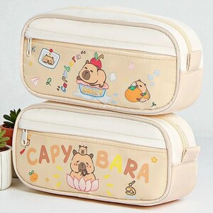 Può includere: Due astucci beige impilati. Ognuno ha una parte superiore bianca e una inferiore beige con cerniera. La parte anteriore di ogni astuccio presenta un disegno di capibara dei cartoni animati con la parola "CAPYBARA".