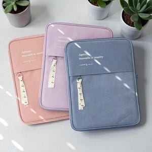 Puede incluir: Tres fundas para tabletas de colores pastel: rosa, lavanda y azul. Cada funda tiene una cremallera, un tirador decorativo y el texto "Apricity, trouvaille in usuality". Las fundas son rectangulares con esquinas redondeadas.