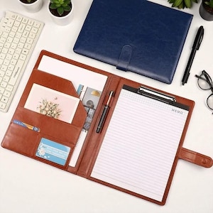 Può includere: Una cartella in pelle marrone con più tasche, un blocco note e una penna. Un taccuino in pelle blu è chiuso sullo sfondo. Sono visibili anche una tastiera, delle piante e degli occhiali.