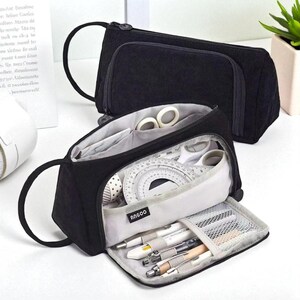 May include: Two black corduroy pencil cases, one open to display stationery. The open case reveals pens, pencils, a protractor, scissors, and tape. The cases have a zippered closure and a handle. The brand name "ANGOO" is visible on the open case.