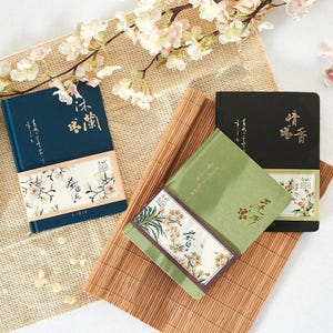 Può includere: Tre quaderni con copertina rigida in verde acqua, verde e nero, ciascuno con disegni floreali e caratteri asiatici. I quaderni sono disposti su un tappetino di bambù e un tessuto beige, con rami di fiori di ciliegio sullo sfondo.