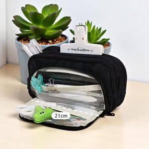 Puede incluir: Estuche negro con cremallera y bolsillo de malla transparente, abierto para revelar bolígrafos, clips y otros artículos de papelería. Un clip blanco con el texto "Near & uniform" está en la parte superior. El estuche abierto mide 21 cm.