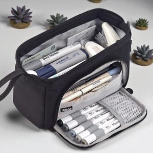 Peut inclure: Trousse à crayons rectangulaire noire avec fermeture éclair, ouverte pour révéler divers articles de papeterie. À l'intérieur, des stylos, des marqueurs, une carte-cadeau Starbucks et une gomme blanche. Sur une surface blanche, avec des plantes succulentes en arrière-plan.