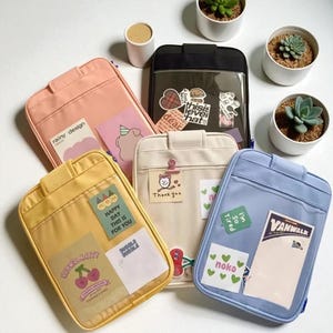 Puede incluir: Una variedad de estuches con cremallera de colores en tonos rosa, amarillo, beige, negro y azul. Cada estuche presenta un bolsillo frontal transparente con pegatinas y notas decorativas. Los estuches están dispuestos sobre una superficie blanca, con pequeñas plantas en macetas en el fondo.