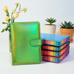 May include: A green iridescent planner with a snap closure stands next to a stack of rainbow-edged notebooks. The notebooks have blue, black, red, and purple covers. A vase of yellow flowers and potted succulents are in the background.