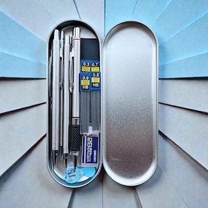 May include: A silver metal pencil case, open to reveal several silver mechanical pencils, lead refills, a blue pencil sharpener, and an eraser. The lead refills are labeled with sizes 0.3, 0.7, 0.9, and 0.5, and the eraser is labeled "2B Eraser".