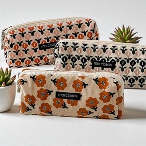 Puede incluir: Tres estuches de lápices de pana con estampados florales en naranja, rosa y negro sobre un fondo crema. Cada estuche tiene cierre de cremallera y una etiqueta "merciparis". Una pequeña planta suculenta es visible en el fondo.