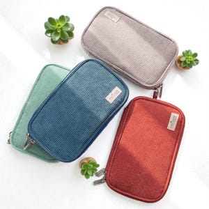 May include: Four corduroy zippered pouches in green, blue, beige, and red. Each pouch has a rectangular shape with a vertical ribbed texture and a small label. Small potted succulents are placed around the pouches.