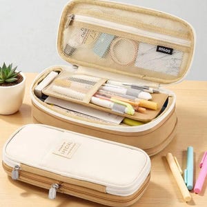 May include: Beige and tan pencil case with a zip closure. The case is open, revealing pens, pencils, and other stationery items. The top compartment has a mesh pocket. The closed case has the word "SPECIAL" on the front.