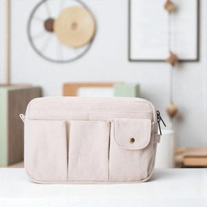 Puede incluir: Un bolso rectangular beige claro con múltiples bolsillos delanteros y un bolsillo pequeño con un botón dorado. El bolso tiene cierre de cremallera y un asa lateral. El bolso está hecho de un material suave y texturizado.