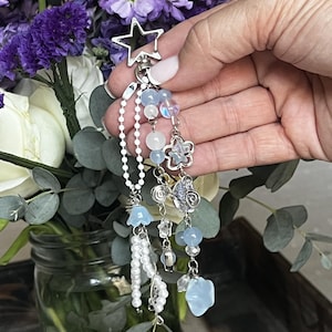 May include: A silver-toned keychain with a star-shaped top and dangling charms. The charms include light blue beads, a flower, and a pearlescent chain. The background features a bouquet of purple and white flowers.