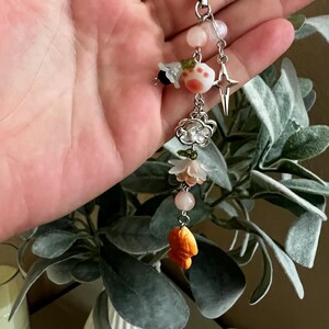 Peut inclure: Un charm de perles avec un cordon blanc, comprenant divers éléments décoratifs. Ceux-ci incluent des perles roses et blanches, un charm en forme d'étoile, un charm en forme de nuage et un petit poisson orange. Le charm est tenu dans une main.