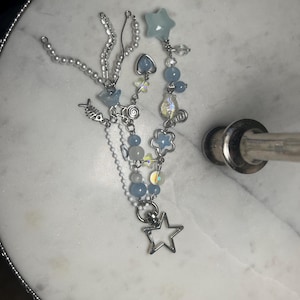 May include: A handmade beaded necklace with a variety of charms and beads. The necklace features light blue, clear, and iridescent beads, along with silver-toned charms including a star, fish, and flower. The necklace is displayed on a white marble surface.