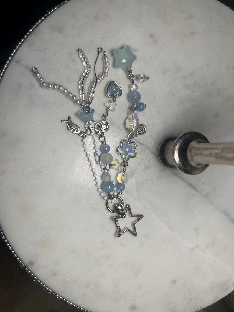 May include: A handmade beaded necklace with a variety of charms and beads. The necklace features light blue, clear, and iridescent beads, along with silver-toned charms including a star, fish, and flower. The necklace is displayed on a white marble surface.