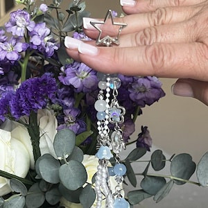 May include: A silver star-shaped ring with a dangling charm featuring beads and pearls. The charm includes light blue and clear beads, small flower charms, and a heart-shaped bead. The ring is held in front of a bouquet of purple and white flowers.