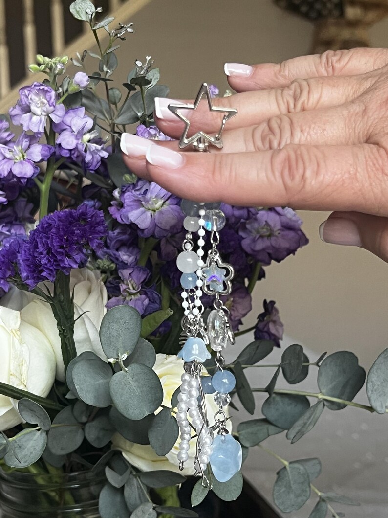 May include: A silver star-shaped ring with a dangling charm featuring beads and pearls. The charm includes light blue and clear beads, small flower charms, and a heart-shaped bead. The ring is held in front of a bouquet of purple and white flowers.