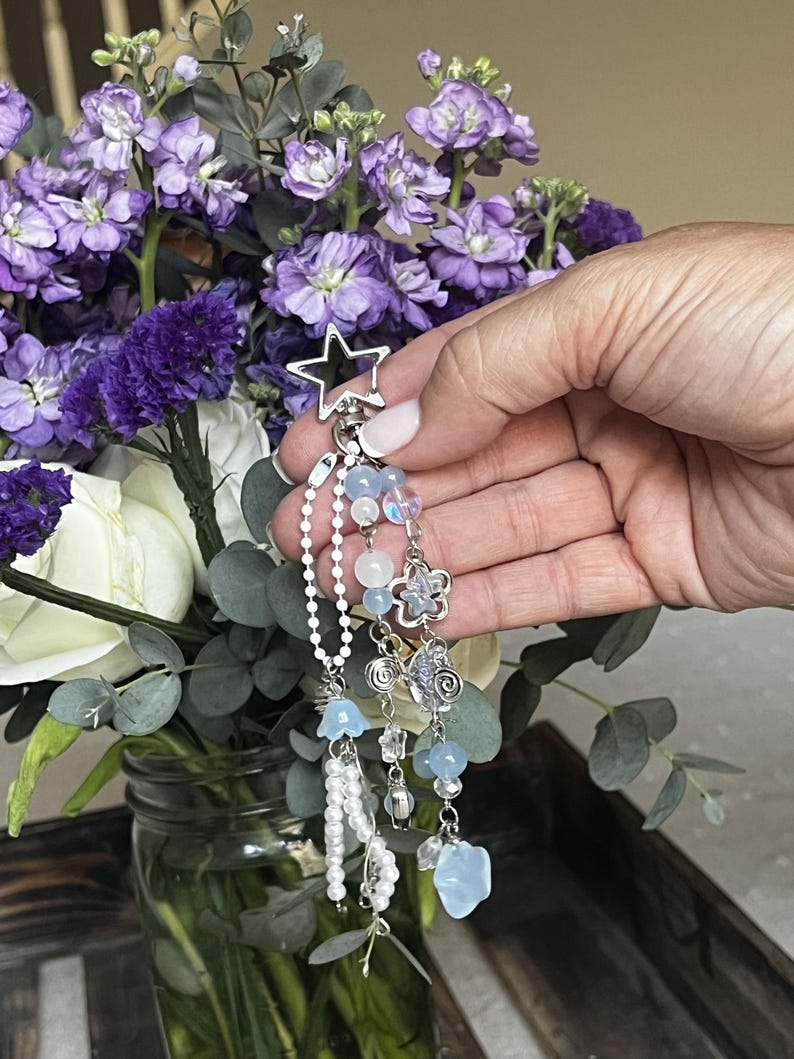 May include: A silver-toned keychain with a star-shaped top and dangling charms. The charms include light blue beads, a flower, and a pearlescent chain. The background features a bouquet of purple and white flowers.