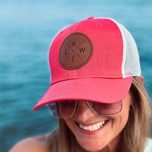 Może przedstawiać: Różowa czapka typu trucker z białą siatką z tyłu i brązową skórzaną naszywką. Na naszywce litery "K", "L" i "W" ułożone wokół krzyża. Czapkę nosi osoba w okularach przeciwsłonecznych.