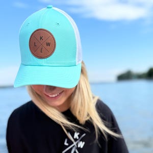 Peut inclure: Casquette de camionneur turquoise et blanche avec un écusson en cuir marron portant les lettres "K", "L" et "W" dans un motif en croix. Le chapeau est associé à un t-shirt noir avec le même logo. Le fond montre un ciel bleu et de l'eau.