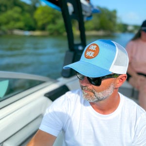 May include: A light blue trucker hat with a white mesh back and a circular patch that reads "Lake Waco". The hat is worn by a person wearing sunglasses and a white t-shirt with a compass design.