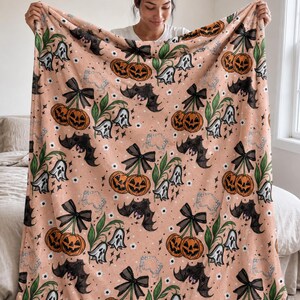 Op de afbeelding: Een zachte perzikkleurige fleece deken met een herhalend patroon van pompoenen gestileerd als kersen met zwarte strikken, witte spookfiguren, donkergrijze vleermuizen, groene bladeren en kleine witte bloemetjes verspreid over de stof.