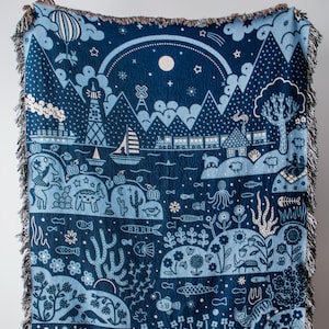 May include: A woven throw blanket in shades of dark blue and light blue featuring a detailed landscape illustration. The design includes a lighthouse, a train, mountains, a sailboat, an octopus, various fish, flowers, and a person gardening.