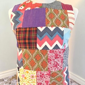 May include: A sleeveless patchwork dress with a variety of fabric patterns, including floral, plaid, and geometric designs. The dress features a mix of colors, such as red, purple, orange, and blue. The dress is displayed on a white mannequin.