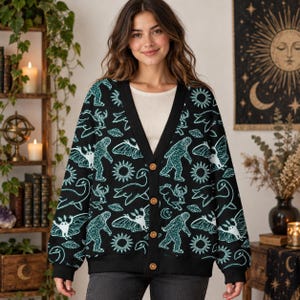 May include: A black V-neck cardigan featuring a repeating pattern of teal-colored cryptids, including Bigfoot, the Loch Ness Monster, and moth-like creatures, along with suns and UFOs. The garment has a button-front closure with four visible brown buttons.
