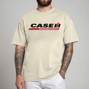 May include: Cream-colored t-shirt featuring the word "CASE" in black, with a red stripe and the word "II" in red. Below the logo, the text reads "DEERE BELONG ON YOUR WALL". The shirt has short sleeves and a crew neck.